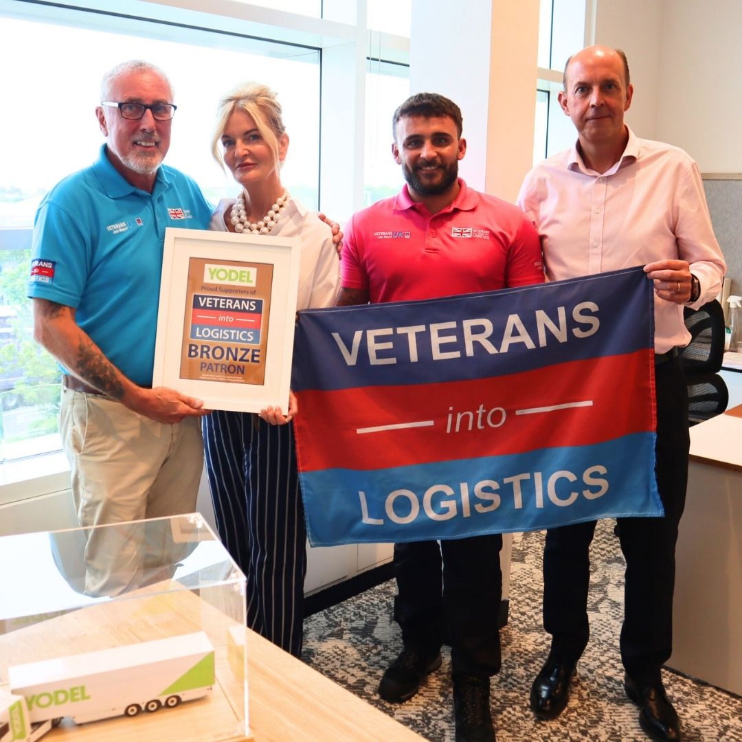 Four people standing in an office, holding a framed certificate and a "Veterans into Logistics" flag.
